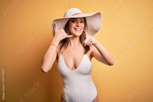 Young beautiful brunette woman on vacation wearing swimsuit and summer hat smiling doing talking on the telephone gesture and pointing to you. Call me.