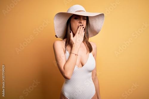 Young beautiful brunette woman on vacation wearing swimsuit and summer hat bored yawning tired covering mouth with hand. Restless and sleepiness.