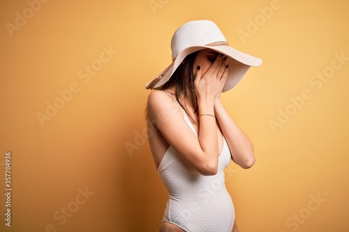 Young beautiful brunette woman on vacation wearing swimsuit and summer hat with sad expression covering face with hands while crying. Depression concept.