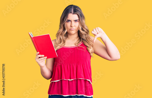 Young caucasian woman holding book with angry face  negative sign showing dislike with thumbs down  rejection concept