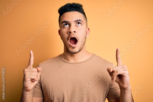 Young handsome man wearing casual t-shirt standing over isolated yellow background amazed and surprised looking up and pointing with fingers and raised arms.