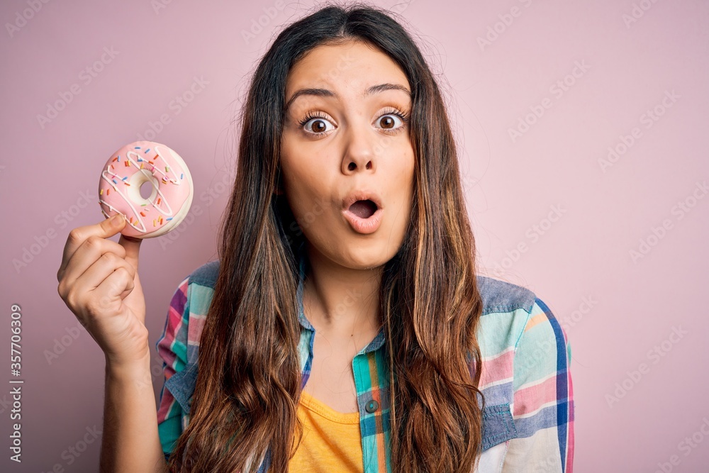 Young beautiful brunette woman eating sweet pink doughnut over isolated background scared in shock with a surprise face, afraid and excited with fear expression Young beautiful brunette woman eating sweet pink doughnut over isolated background scared in shock with a surprise face, afraid and excited with fear expression