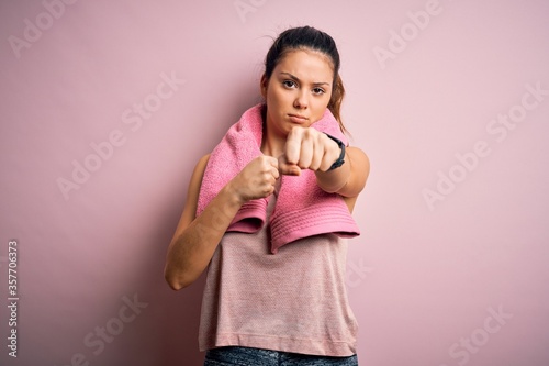 Young beautiful brunette sportswoman wearing sportswear and towel over pink background Punching fist to fight, aggressive and angry attack, threat and violence