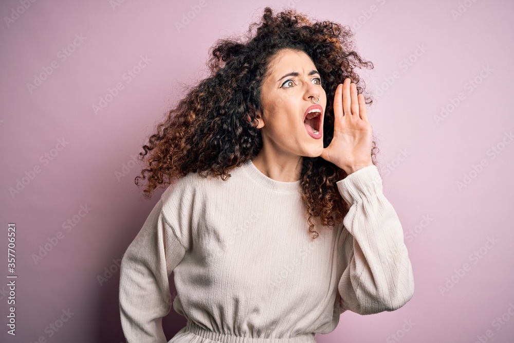 Beautiful woman with curly hair and piercing wearing casual sweater over pink background shouting and screaming loud to side with hand on mouth. Communication concept.