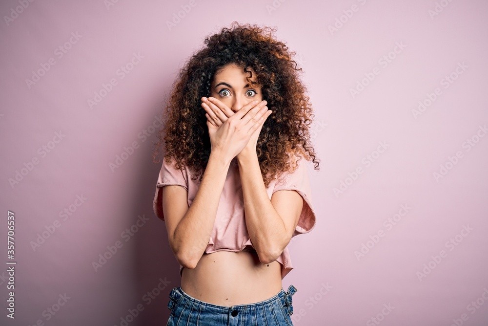 Young beautiful woman with curly hair and piercing wearing casual pink t-shirt shocked covering mouth with hands for mistake. Secret concept.