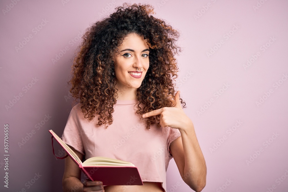 Young beautiful student woman with curly hair and piercing reading book over pink background with surprise face pointing finger to himself Young beautiful student woman with curly hair and piercing reading book over pink background with surprise face pointing finger to himself