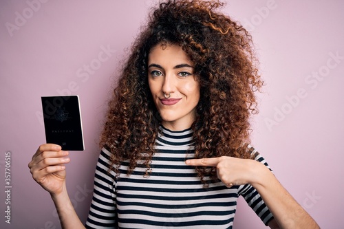 Beautiful tourist woman with curly hair and piercing holding australia australian passport id with surprise face pointing finger to himself