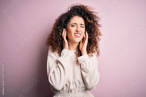 Beautiful woman with curly hair and piercing wearing casual sweater over pink background covering ears with fingers with annoyed expression for the noise of loud music. Deaf concept. © Krakenimages.com