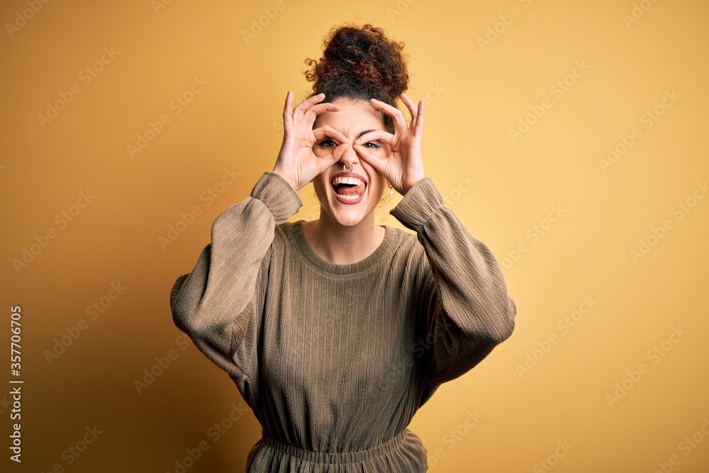 Young beautiful brunette woman with curly hair and piercing wearing casual dress doing ok gesture like binoculars sticking tongue out, eyes looking through fingers. Crazy expression. Young beautiful brunette woman with curly hair and piercing wearing casual dress doing ok gesture like binoculars sticking tongue out, eyes looking through fingers. Crazy expression.