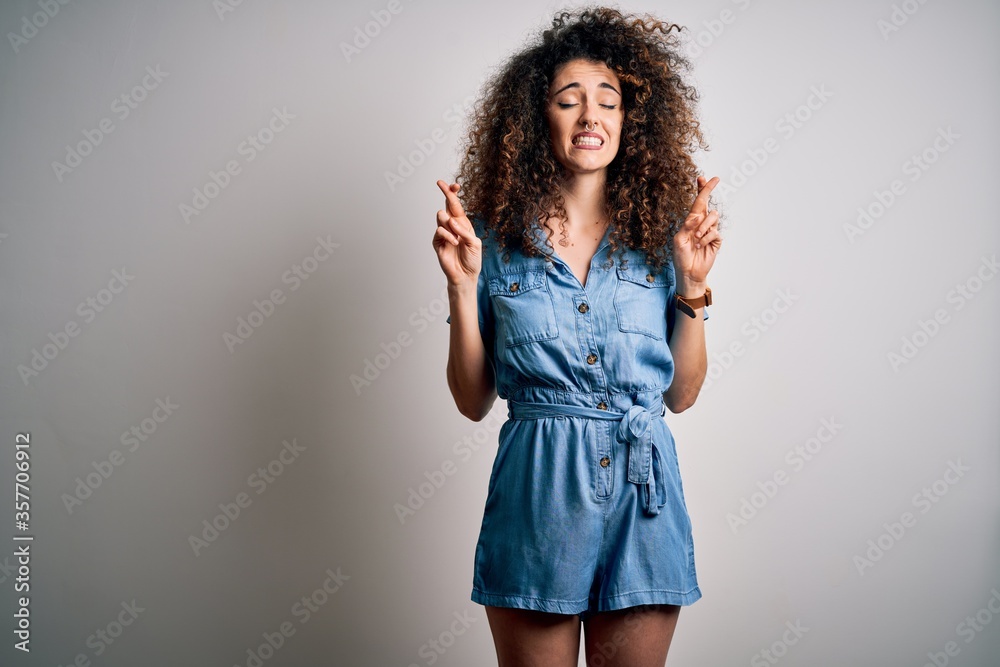 Young beautiful woman with curly hair and piercing wearing casual denim dress gesturing finger crossed smiling with hope and eyes closed. Luck and superstitious concept. Young beautiful woman with curly hair and piercing wearing casual denim dress gesturing finger crossed smiling with hope and eyes closed. Luck and superstitious concept.
