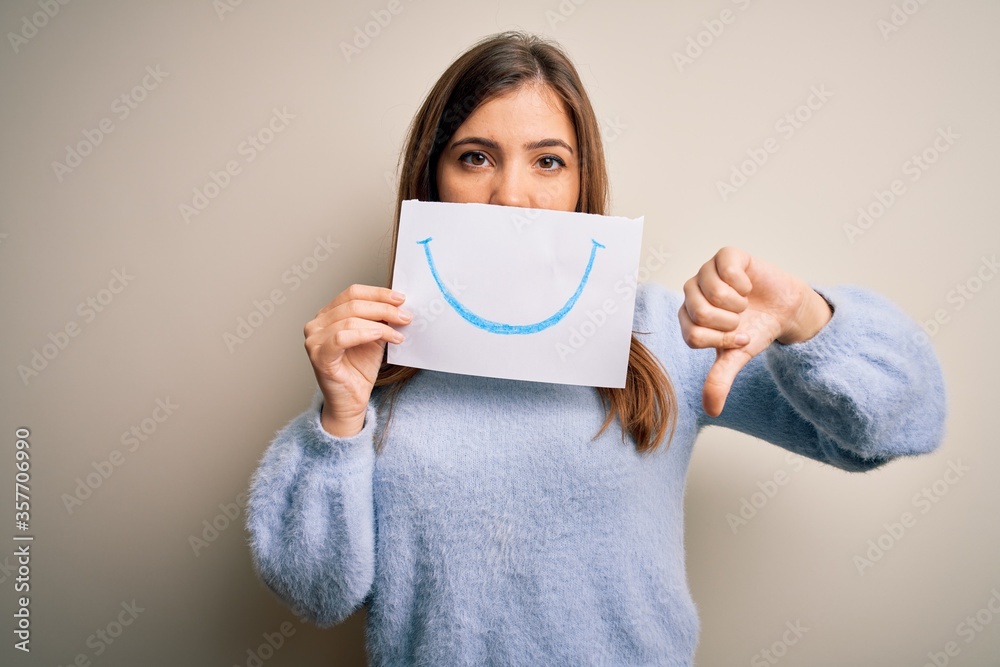 Young blonde woman holding funny smile drawing on mouth as happy expression with angry face, negative sign showing dislike with thumbs down, rejection concept Young blonde woman holding funny smile drawing on mouth as happy expression with angry face, negative sign showing dislike with thumbs down, rejection concept