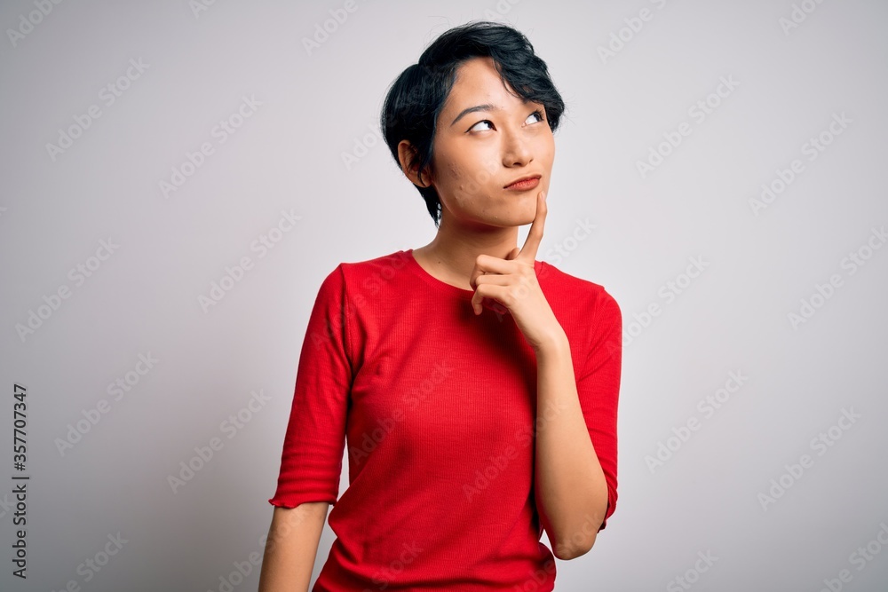 Young beautiful asian girl wearing casual red t-shirt standing over isolated white background with hand on chin thinking about question, pensive expression. Smiling and thoughtful face. Doubt concept.