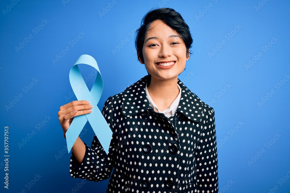 Young beautiful asian girl holding blue cancer ribbon symbol over isolated background with a happy face standing and smiling with a confident smile showing teeth Young beautiful asian girl holding blue cancer ribbon symbol over isolated background with a happy face standing and smiling with a confident smile showing teeth