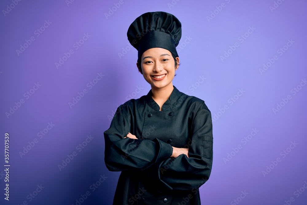 Young beautiful chinese chef woman wearing cooker uniform and hat over purple background happy face smiling with crossed arms looking at the camera. Positive person. Young beautiful chinese chef woman wearing cooker uniform and hat over purple background happy face smiling with crossed arms looking at the camera. Positive person.