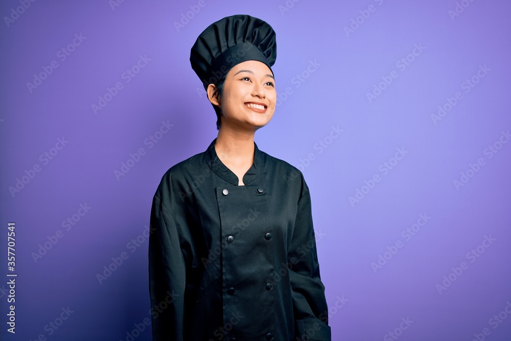 Young beautiful chinese chef woman wearing cooker uniform and hat over purple background looking away to side with smile on face, natural expression. Laughing confident. Young beautiful chinese chef woman wearing cooker uniform and hat over purple background looking away to side with smile on face, natural expression. Laughing confident.