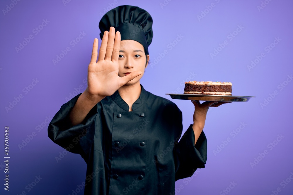 Young chinese chef woman wearing cooker uniform and hat holding tray with cake with open hand doing stop sign with serious and confident expression, defense gesture Young chinese chef woman wearing cooker uniform and hat holding tray with cake with open hand doing stop sign with serious and confident expression, defense gesture