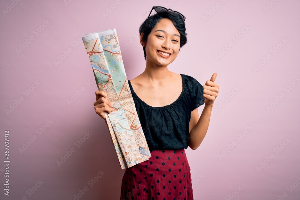 Young beautiful asian tourist girl on vacation holding city map over isolated pink background happy with big smile doing ok sign, thumb up with fingers, excellent sign Young beautiful asian tourist girl on vacation holding city map over isolated pink background happy with big smile doing ok sign, thumb up with fingers, excellent sign