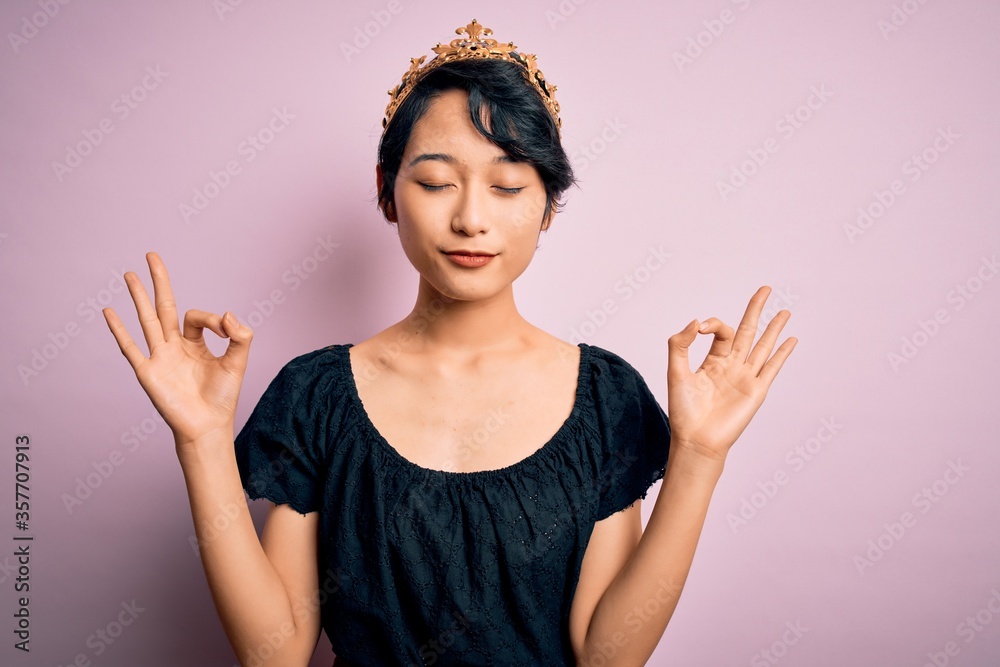 Young beautiful chinese woman wearing golden crown of king over isolated pink background relax and smiling with eyes closed doing meditation gesture with fingers. Yoga concept. Young beautiful chinese woman wearing golden crown of king over isolated pink background relax and smiling with eyes closed doing meditation gesture with fingers. Yoga concept.