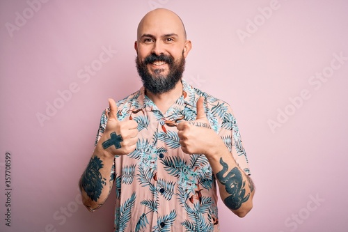 Handsome bald man with beard and tattoo wearing casual floral shirt over pink background success sign doing positive gesture with hand, thumbs up smiling and happy. Cheerful expression.