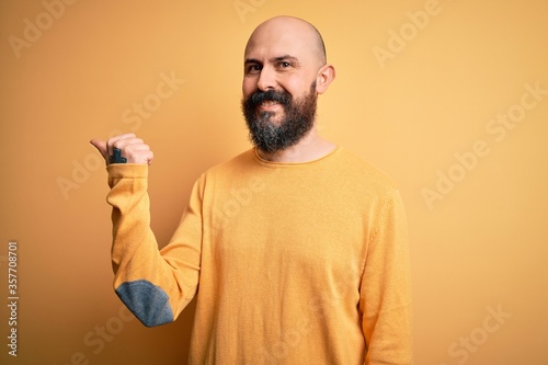 Handsome bald man with beard wearing casual sweater standing over yellow background smiling with happy face looking and pointing to the side with thumb up.