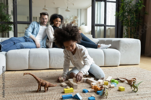 African daughter play on floor with wooden blocks set and dinosaur toys while multi ethnic parents sitting on sofa in living room, homeowners spend time at new home. First house family weekend concept