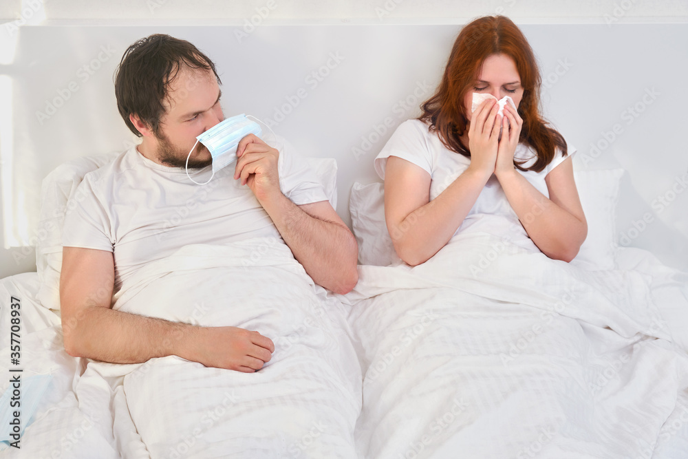 A man is protected by a medical mask from a sick woman lying together in a white bed, concept. Relationship problems coupled with isolation due to coronavirus.