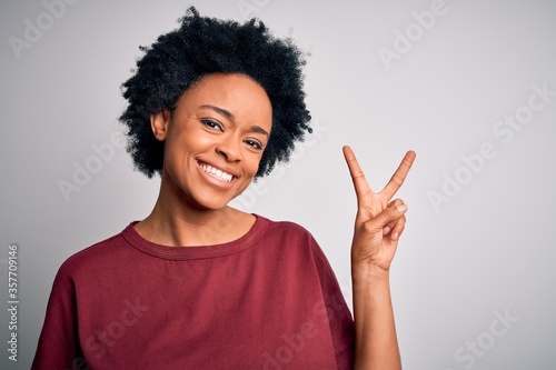 Young beautiful African American afro woman with curly hair wearing casual t-shirt standing smiling with happy face winking at the camera doing victory sign. Number two.