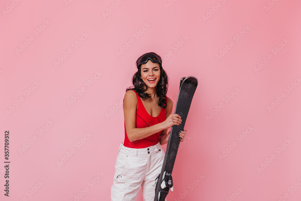 Attractive brunette woman in sport goggles smiling at camera. Magnificent girl with skis standing on pink background.