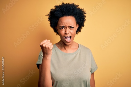 Young beautiful African American afro woman with curly hair wearing casual t-shirt angry and mad raising fist frustrated and furious while shouting with anger. Rage and aggressive concept.