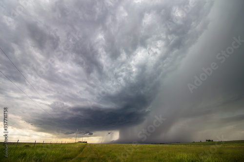 A supercell thunderstorm approaches over the rural countryside, with curtains of rain and hail falling.