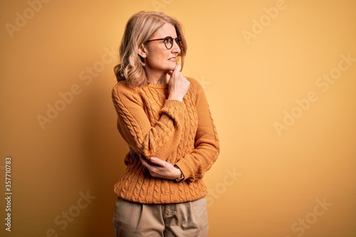 Middle age beautiful blonde woman wearing casual sweater and glasses over yellow background Thinking worried about a question, concerned and nervous with hand on chin © Krakenimages.com