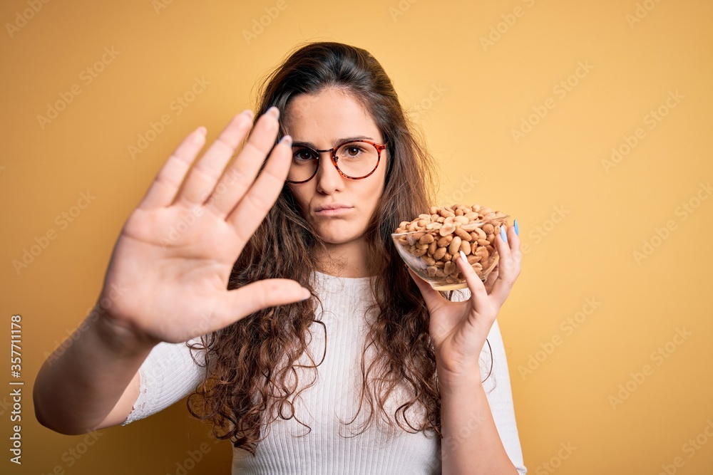 Beautiful woman with curly hair holding bowl with healthy peanuts over yellow background with open hand doing stop sign with serious and confident expression, defense gesture