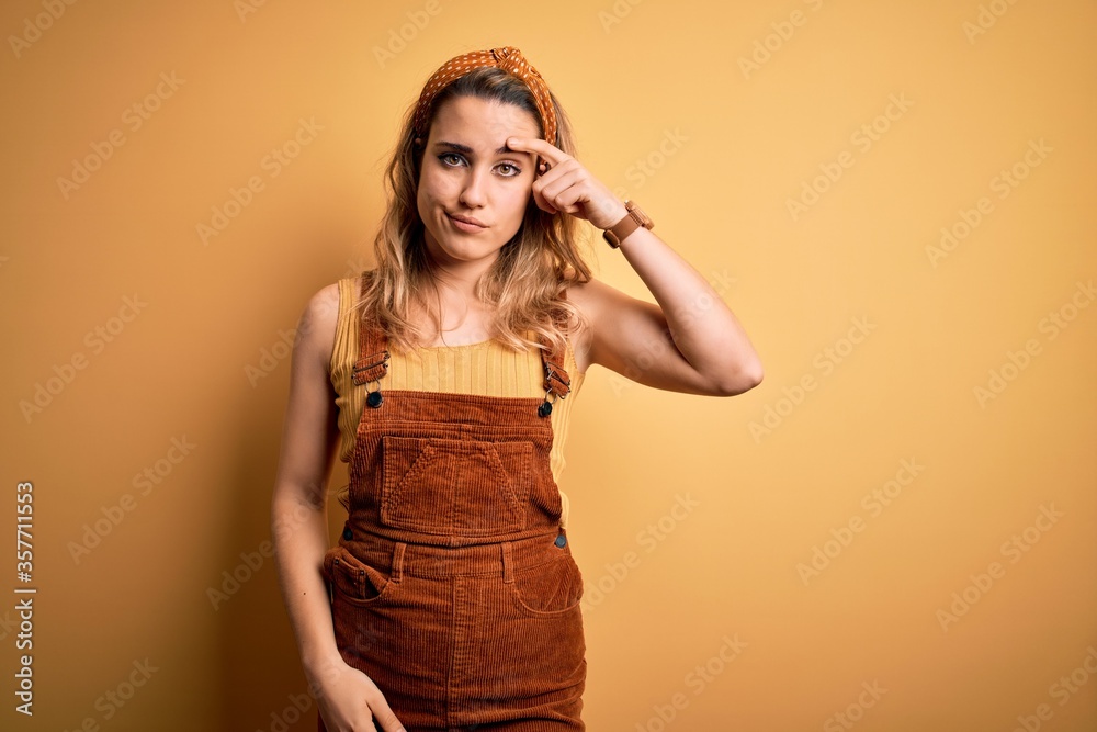 Young beautiful blonde woman wearing overalls and diadem standing over yellow background pointing unhappy to pimple on forehead, ugly infection of blackhead. Acne and skin problem Young beautiful blonde woman wearing overalls and diadem standing over yellow background pointing unhappy to pimple on forehead, ugly infection of blackhead. Acne and skin problem