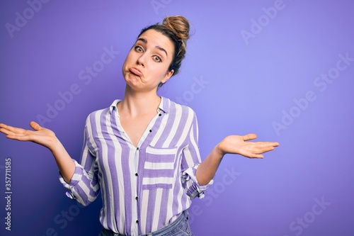 Young beautiful blonde woman wearing casual striped shirt standing over purple background clueless and confused expression with arms and hands raised. Doubt concept.