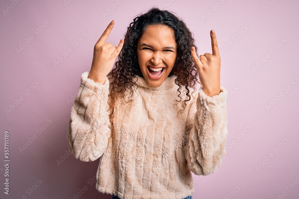 Young beautiful woman with curly hair wearing casual sweater standing over pink background shouting with crazy expression doing rock symbol with hands up. Music star. Heavy music concept. Young beautiful woman with curly hair wearing casual sweater standing over pink background shouting with crazy expression doing rock symbol with hands up. Music star. Heavy music concept.
