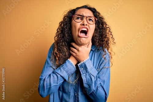 Beautiful woman with curly hair wearing casual denim shirt and glasses over yellow background shouting and suffocate because painful strangle. Health problem. Asphyxiate and suicide concept.