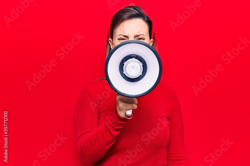 Young beautiful woman wearing diadem with angry expression. Screaming loud using megaphone standing over isolated red background © Krakenimages.com
