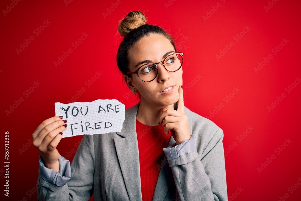 Young beautiful blonde woman with blue eyes holding paper with you are fired message serious face thinking about question, very confused idea Young beautiful blonde woman with blue eyes holding paper with you are fired message serious face thinking about question, very confused idea