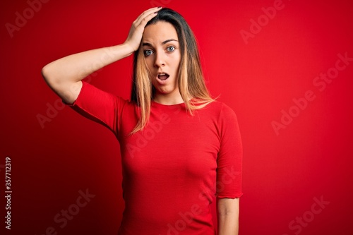 Young beautiful blonde woman with blue eyes wearing casual t-shirt over red background surprised with hand on head for mistake, remember error. Forgot, bad memory concept.