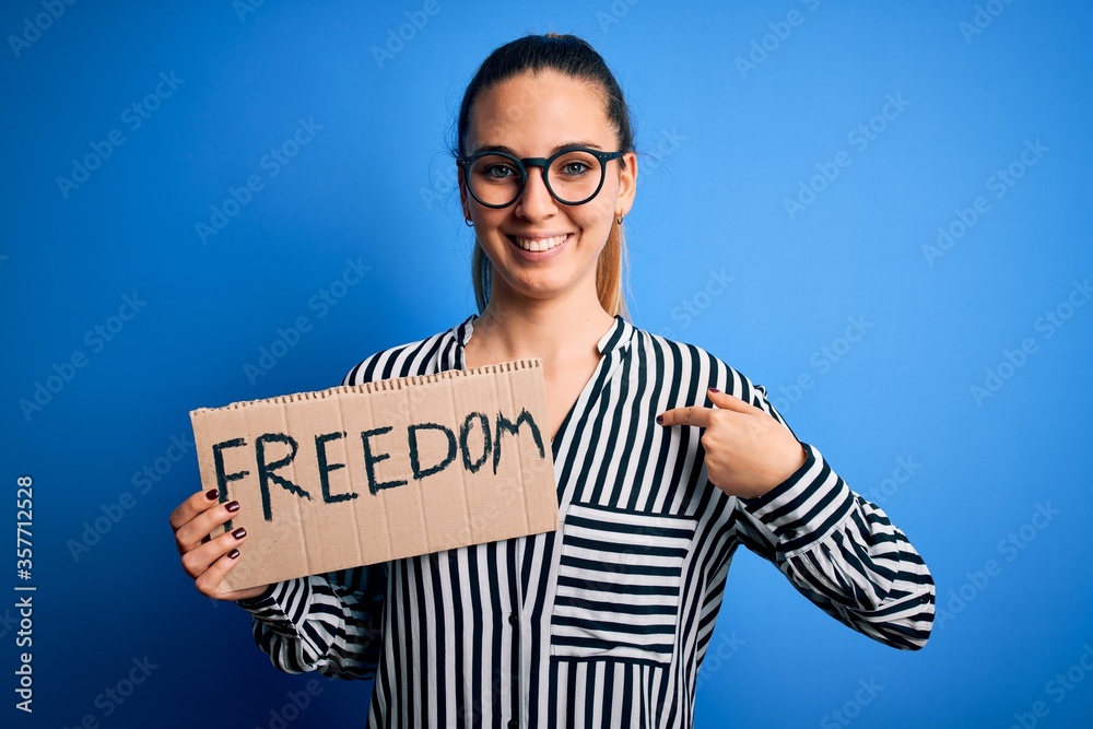 Young beautiful blonde woman with blue eyes holding banner with freedom message with surprise face pointing finger to himself Young beautiful blonde woman with blue eyes holding banner with freedom message with surprise face pointing finger to himself