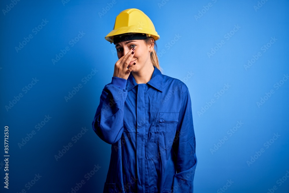 Young beautiful worker woman with blue eyes wearing security helmet and uniform smelling something stinky and disgusting, intolerable smell, holding breath with fingers on nose. Bad smell