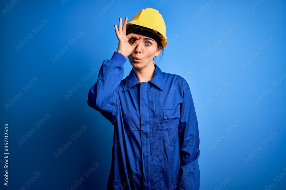 Young beautiful worker woman with blue eyes wearing security helmet and uniform doing ok gesture shocked with surprised face, eye looking through fingers. Unbelieving expression.