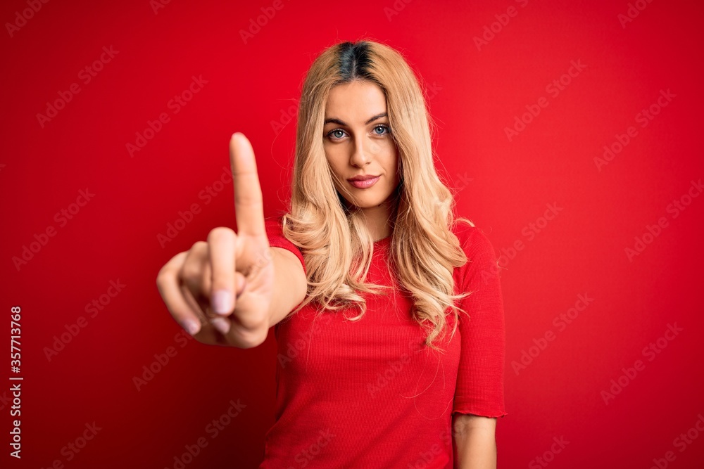 Young beautiful blonde woman wearing casual t-shirt standing over isolated red background Pointing with finger up and angry expression, showing no gesture Young beautiful blonde woman wearing casual t-shirt standing over isolated red background Pointing with finger up and angry expression, showing no gesture
