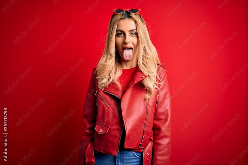 Young beautiful blonde woman wearing casual jacket standing over isolated red background sticking tongue out happy with funny expression. Emotion concept. Young beautiful blonde woman wearing casual jacket standing over isolated red background sticking tongue out happy with funny expression. Emotion concept.