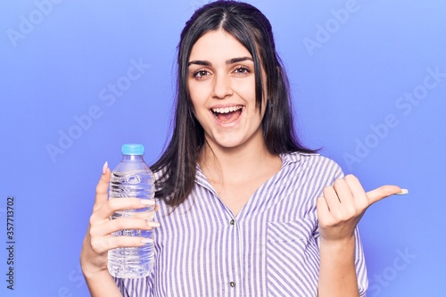 Young beautiful girl holding bottle of water pointing thumb up to the side smiling happy with open mouth