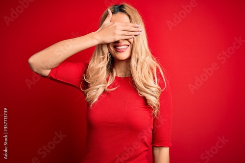 Young beautiful blonde woman wearing casual t-shirt standing over isolated red background smiling and laughing with hand on face covering eyes for surprise. Blind concept.