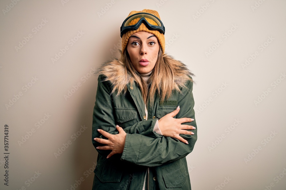 Young brunette skier woman wearing snow clothes and ski goggles over white background shaking and freezing for winter cold with sad and shock expression on face