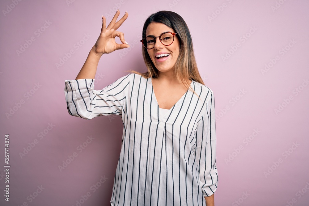 Young beautiful woman wearing casual striped t-shirt and glasses over pink background smiling positive doing ok sign with hand and fingers. Successful expression. Young beautiful woman wearing casual striped t-shirt and glasses over pink background smiling positive doing ok sign with hand and fingers. Successful expression.