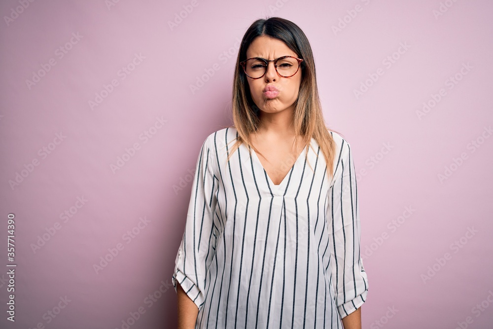 Young beautiful woman wearing casual striped t-shirt and glasses over pink background puffing cheeks with funny face. Mouth inflated with air, crazy expression. Young beautiful woman wearing casual striped t-shirt and glasses over pink background puffing cheeks with funny face. Mouth inflated with air, crazy expression.