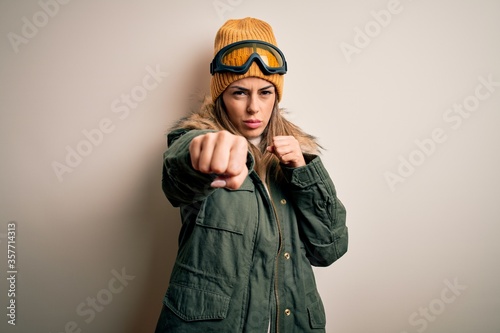Young brunette skier woman wearing snow clothes and ski goggles over white background Punching fist to fight, aggressive and angry attack, threat and violence
